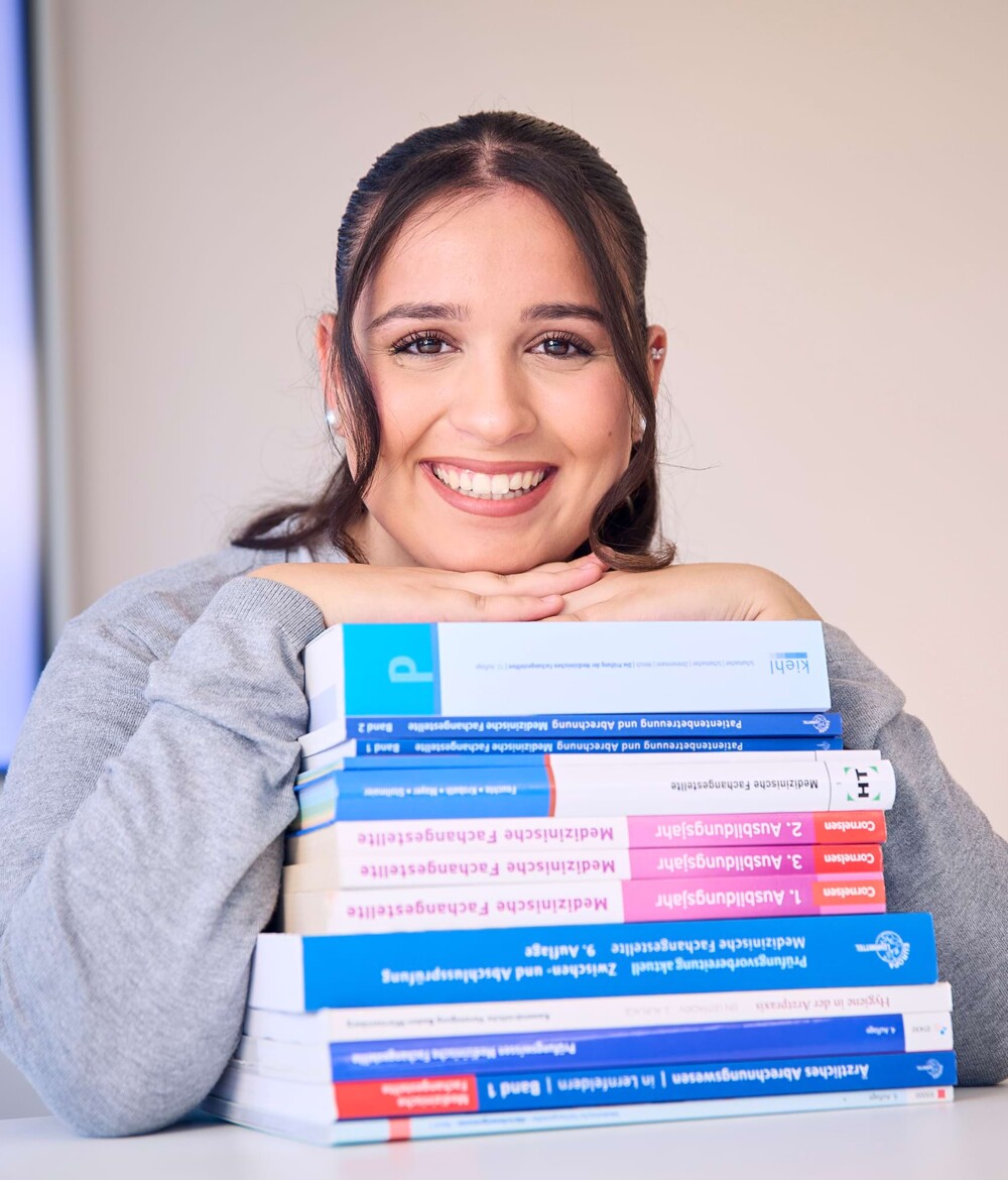 Eine Auszubildende l&auml;chelt in die Kamera und st&uuml;tzt sich auf Lehrb&uuml;chern ab.