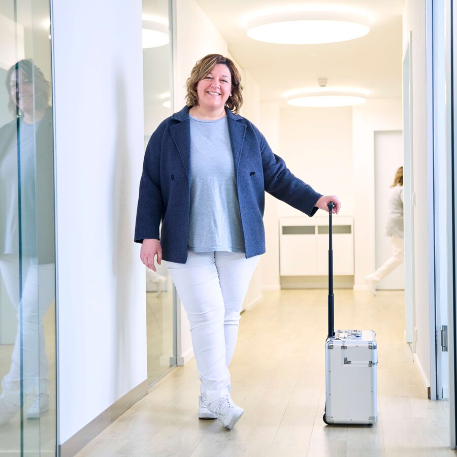 Eine medizinische Fachangestellte steht mit vorbereitetem Koffer im Praxisflur.
