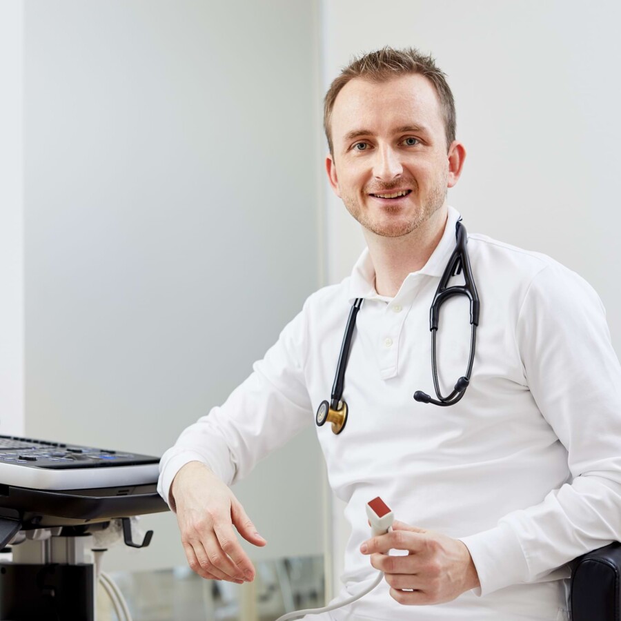 Ein junger Arzt h&auml;lt ein Handmessger&auml;t und l&auml;chelt zur Kamera.