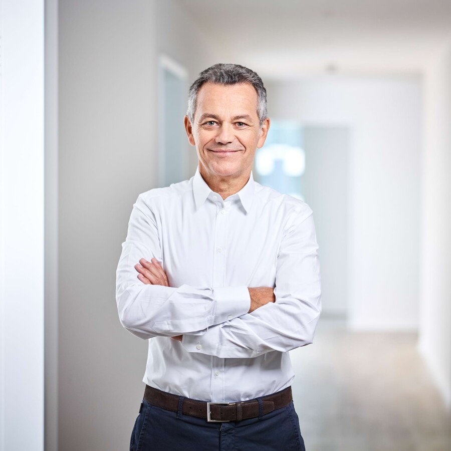 Ein Arzt steht mit verschr&auml;nkten Armen im Flur und l&auml;chelt in die Kamera.