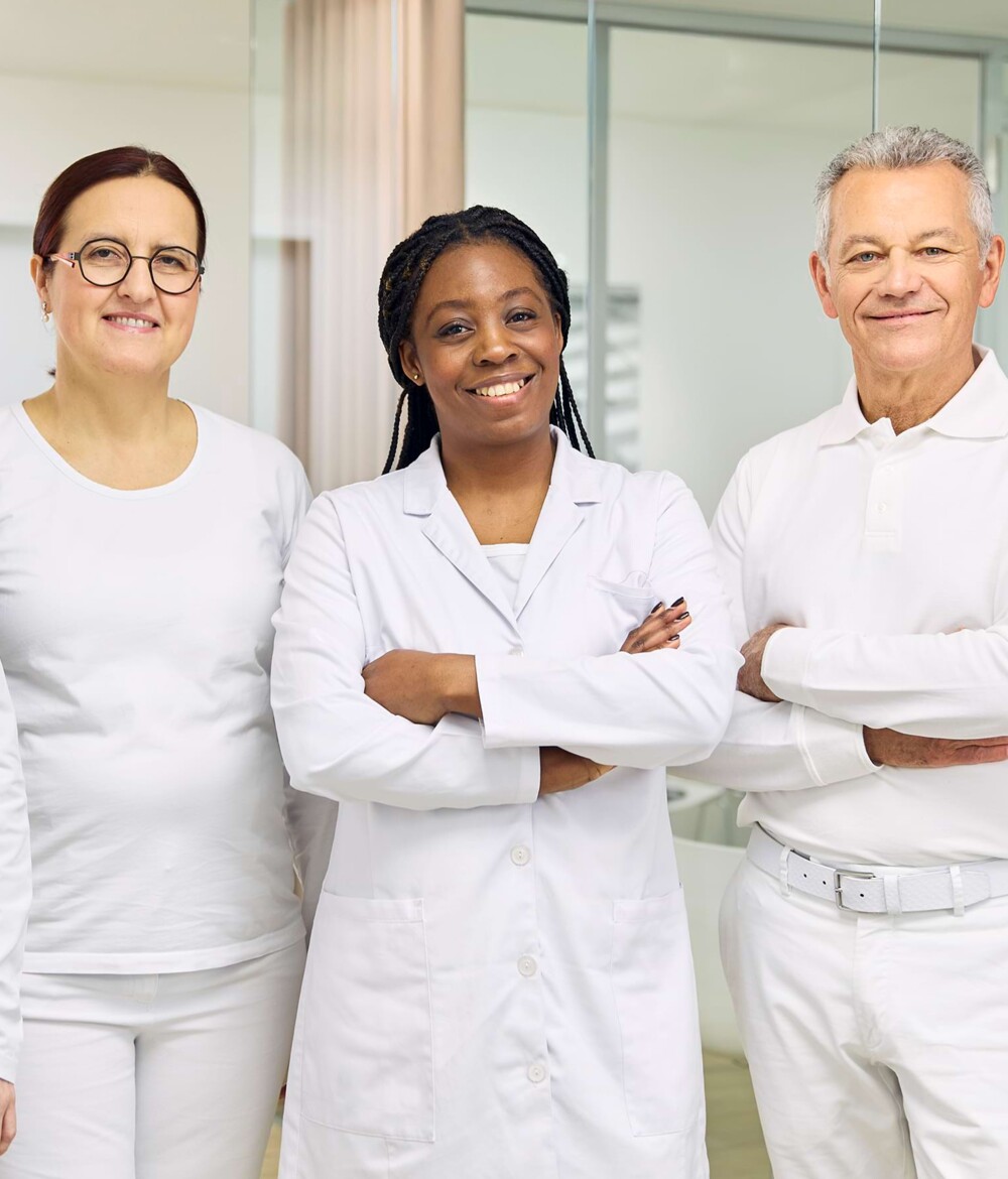 Zwei &Auml;rztinnen und ein Arzt stehen mit verschr&auml;nkten Armen in der Praxis und l&auml;cheln in die Kamera.