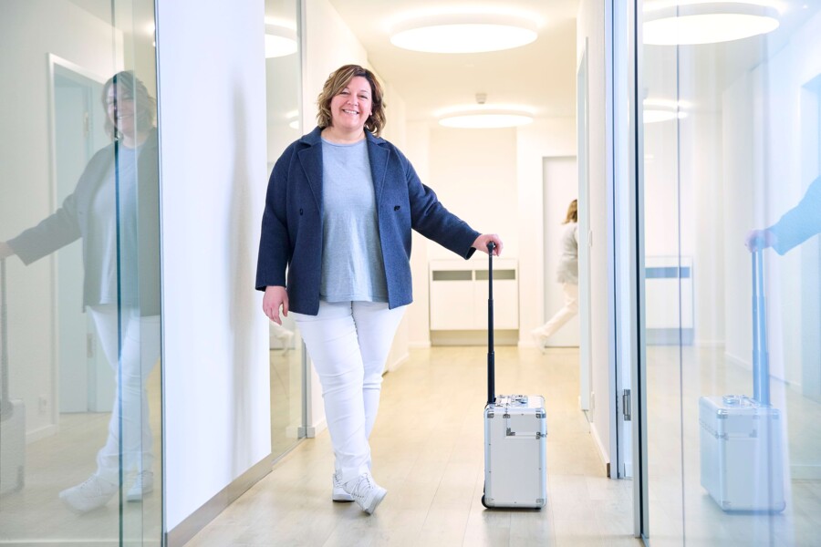 Eine medizinische Fachangestellte steht mit vorbereitetem Koffer im Praxisflur.