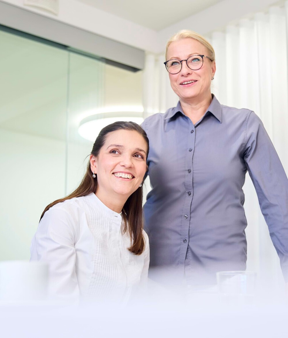 Eine Mitarbeiterin sitzt und schaut lachend an der Kamera vorbei. Neben ihr steht eine weitere Mitarbeiterin.