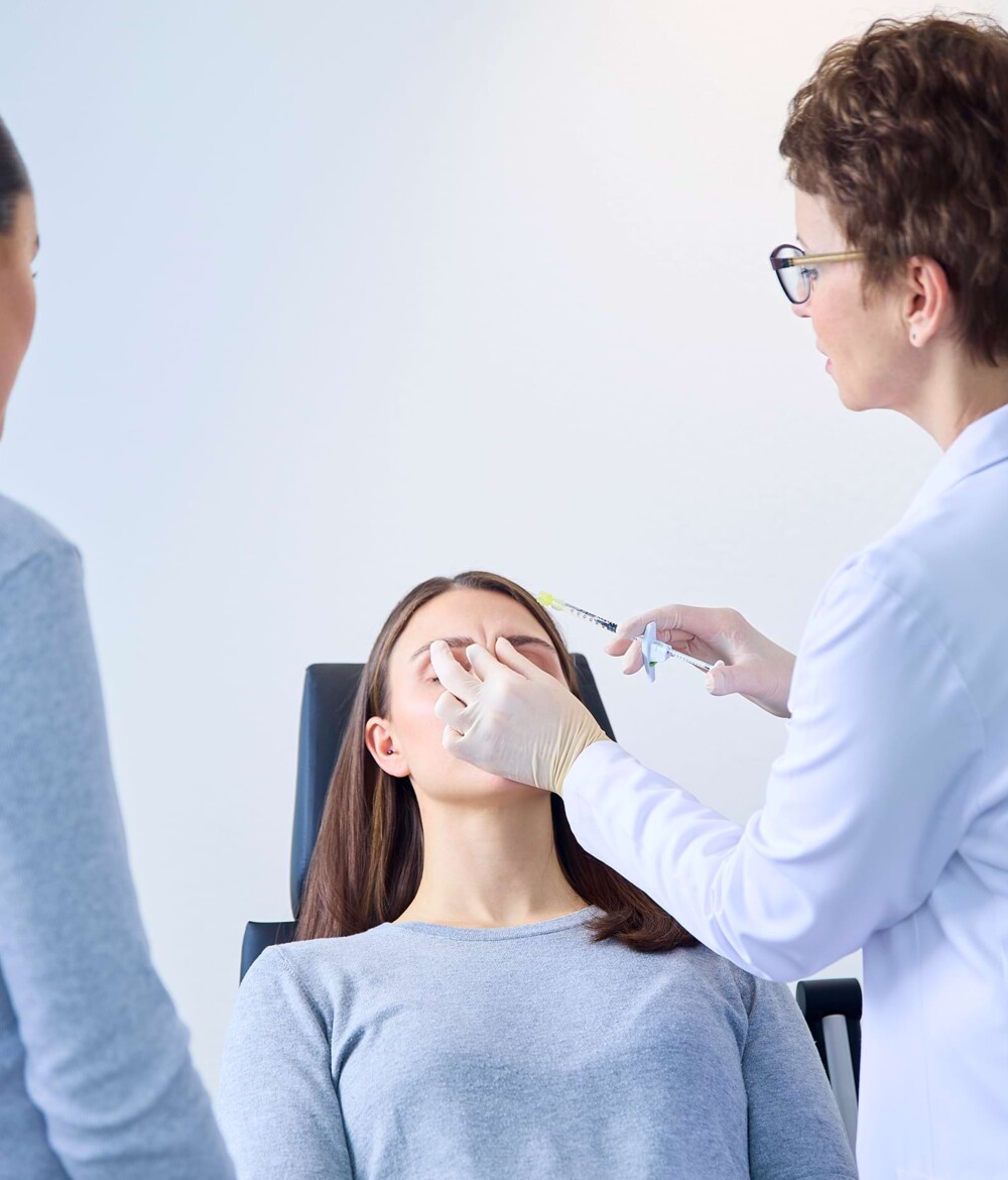 Eine Patientin liegt auf einem Behandlungsstuhl. Momentaufnahme kurz bevor von einer &Auml;rztin in Begleitung einer Arzthelferin eine Botox-Spritze verabreicht wird.