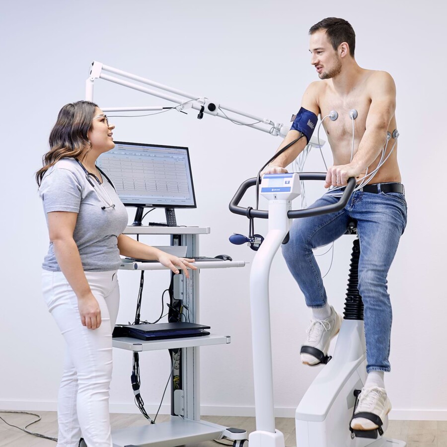 Ein junger Mann sitzt oberk&ouml;rperfrei auf einem Fitnessfahrrad und macht im Rahmen der Sporttauglichkeitsuntersuchung ein Leistungs-EKG.