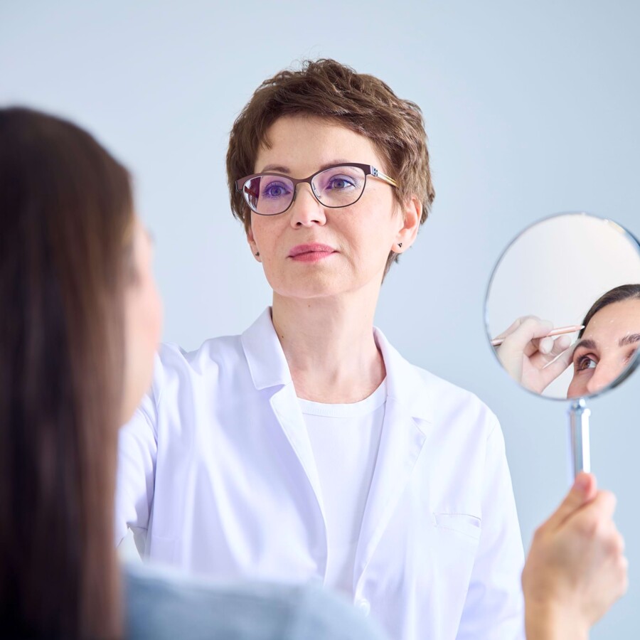 Eine &Auml;rztin h&auml;lt einer Patientin einen Handspiegel vor. Der Spiegel zeigt wie die &Auml;rztin Markierungen im Gesicht der Patientin vornimmt.