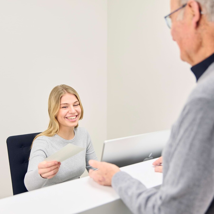 Eine Mitarbeiterin an der Rezeption &uuml;berreicht einem Patienten Unterlagen.
