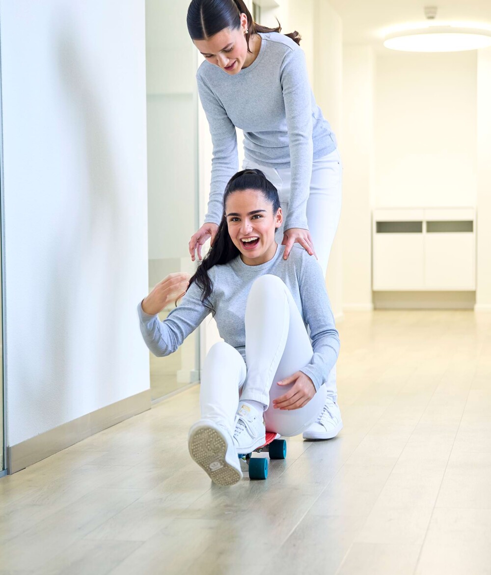 Zwei Mitarbeiterinnen haben Spa&szlig;. Die eine Mitarbeiterin sitzt auf einem Skateboard und wird von ihrer Kollegin lachend durch den Gang geschoben.