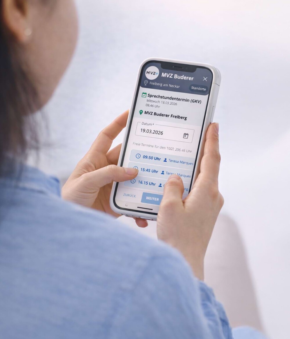 Blick &uuml;ber die Schultern einer jungen Frau mit blauer Bluse und braunen langen Haaren, wie sie ein Smartphone in der Hand h&auml;lt und per Online-Rezeption einen Termin im MVZ Team Buderer bucht