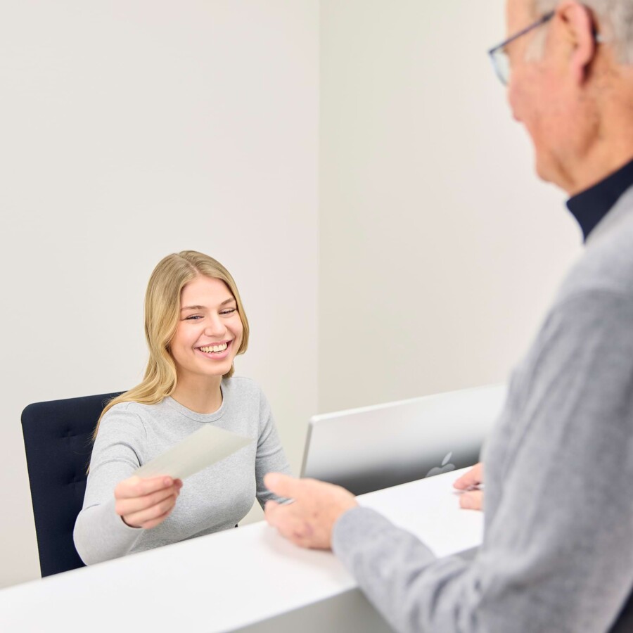 Eine Mitarbeiterin an der Rezeption &uuml;berreicht einem Patienten Unterlagen.