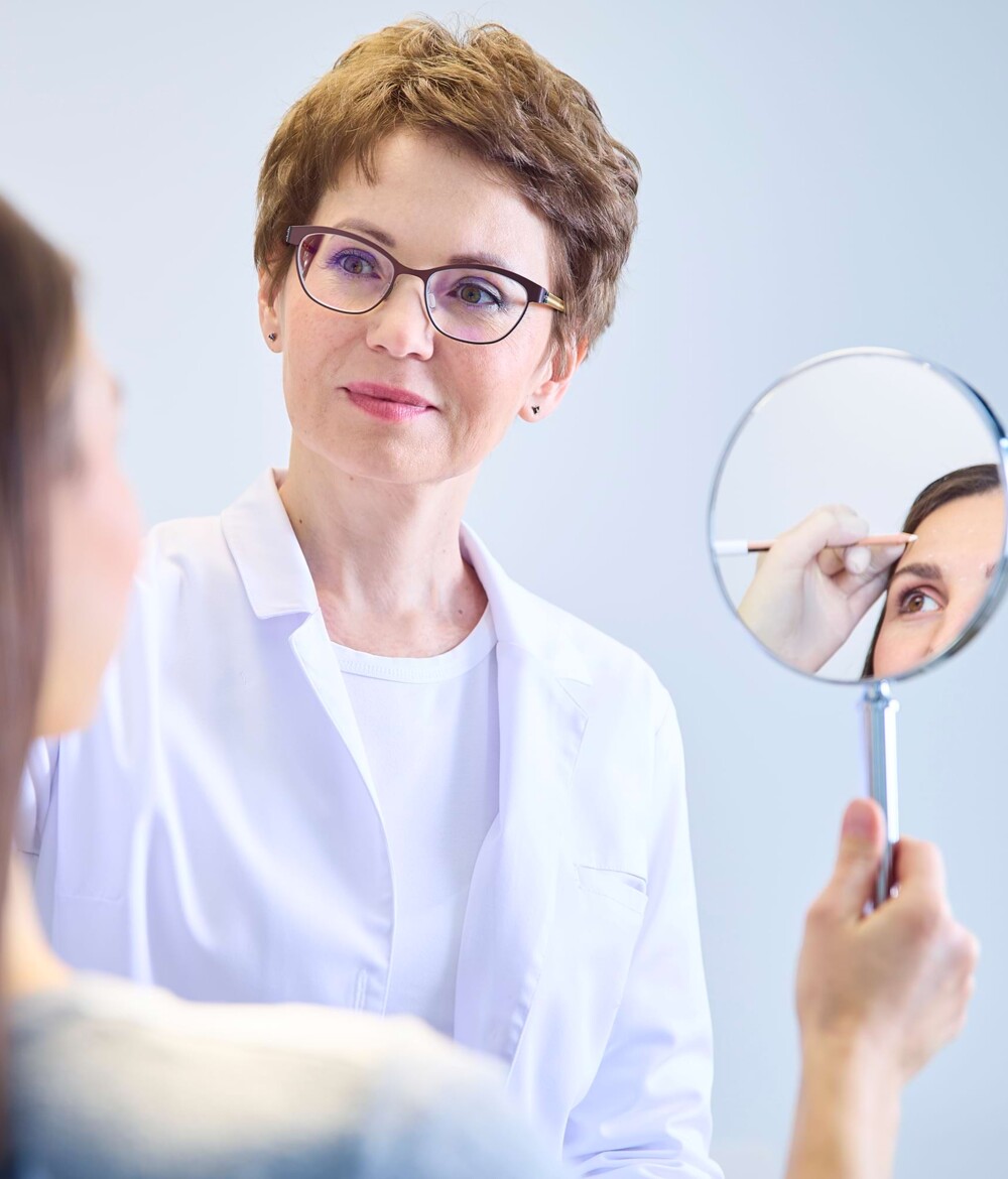 Eine &Auml;rztin h&auml;lt einer Patientin einen Handspiegel vor. Der Spiegel zeigt wie die &Auml;rztin Markierungen im Gesicht der Patientin vornimmt.