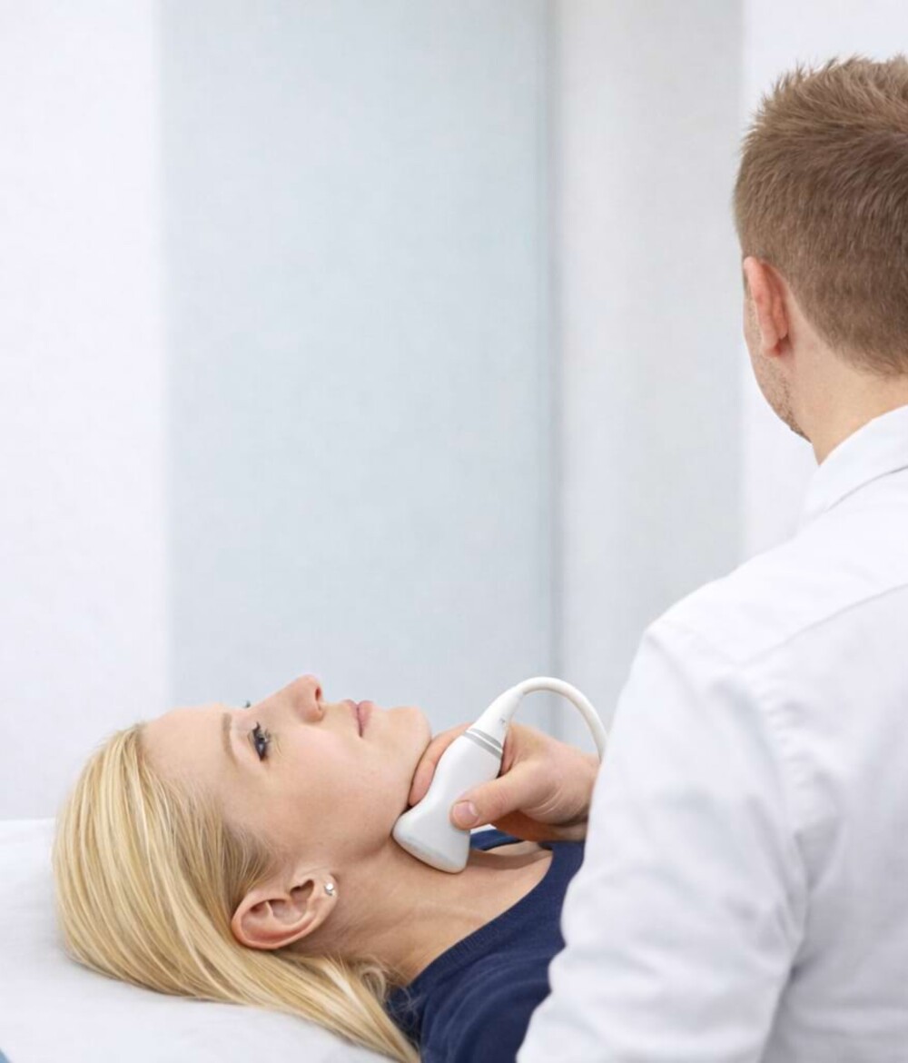 Eine junge Patientin liegt auf einer Behandlungsliege und bekommt von einem Arzt eine Farbduplex-Sonographie der Schlagadern gemacht.